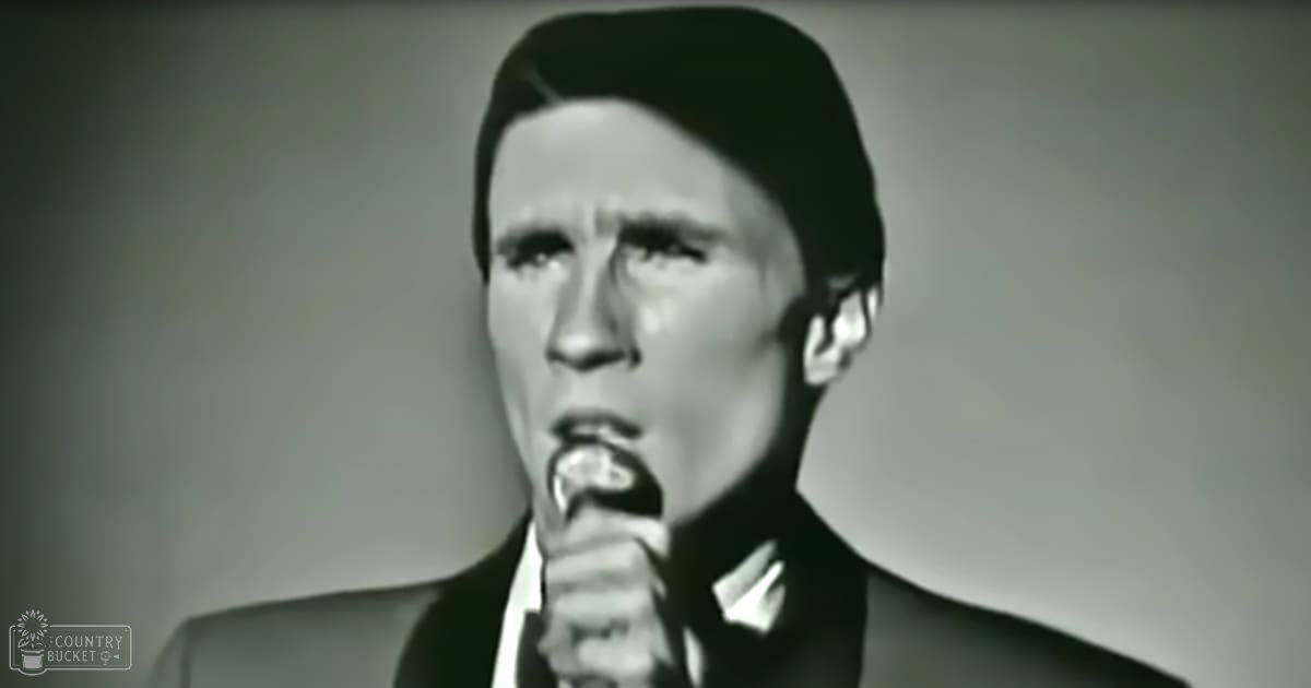 Adult male singer in dark suit jacket with white shirt and tie singing into a microphone on stage.