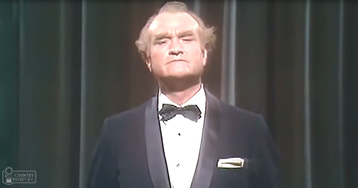 A middle-aged adult male wearing a classic tuxedo with a bow tie, standing indoors on what appears to be a stage with dark curtains in the background.