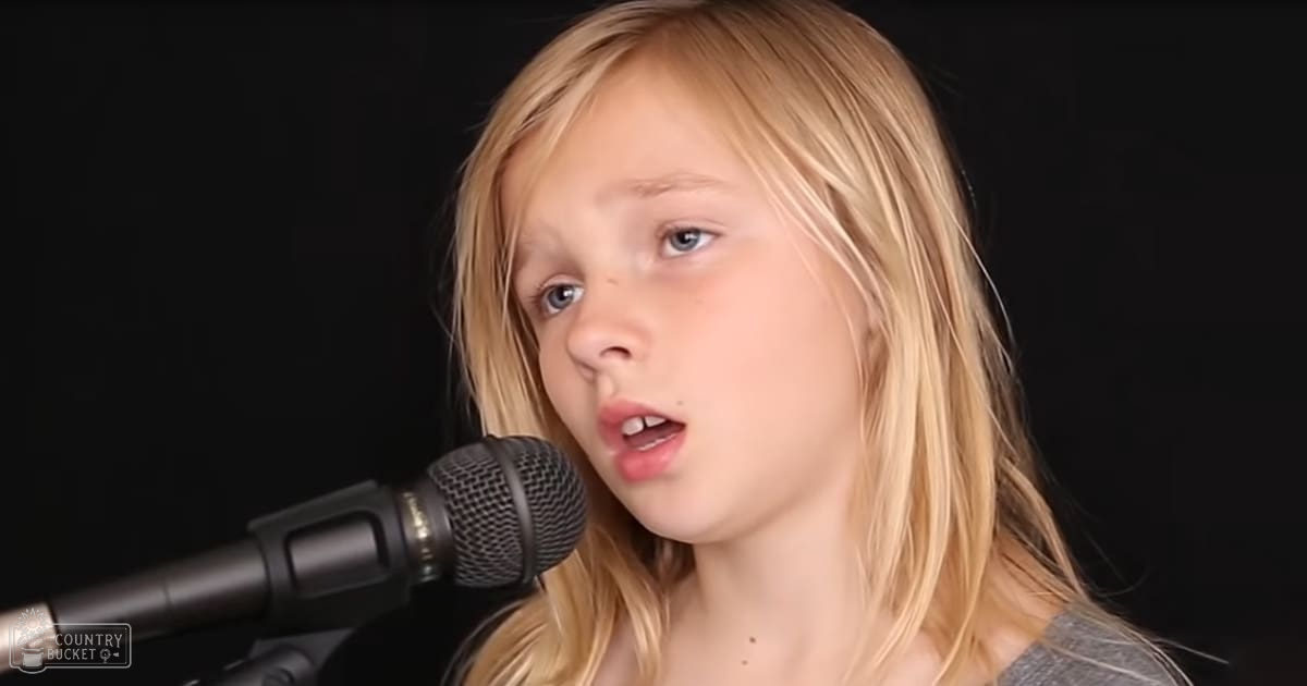 A young female child wearing a gray t-shirt indoors, with a microphone nearby suggesting a recording or performance setup.
