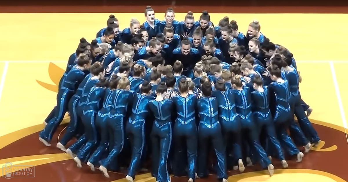 A group of young adult female gymnasts in a huddle wearing matching blue leotards with white trim and patterns, standing on an indoor sports facility floor designed to resemble a basketball or gymnastics court.