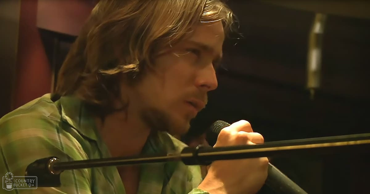 A young adult male in a recording studio, wearing a green and white striped shirt from the 1980s or 1990s, surrounded by soundproofing panels and equipment racks.