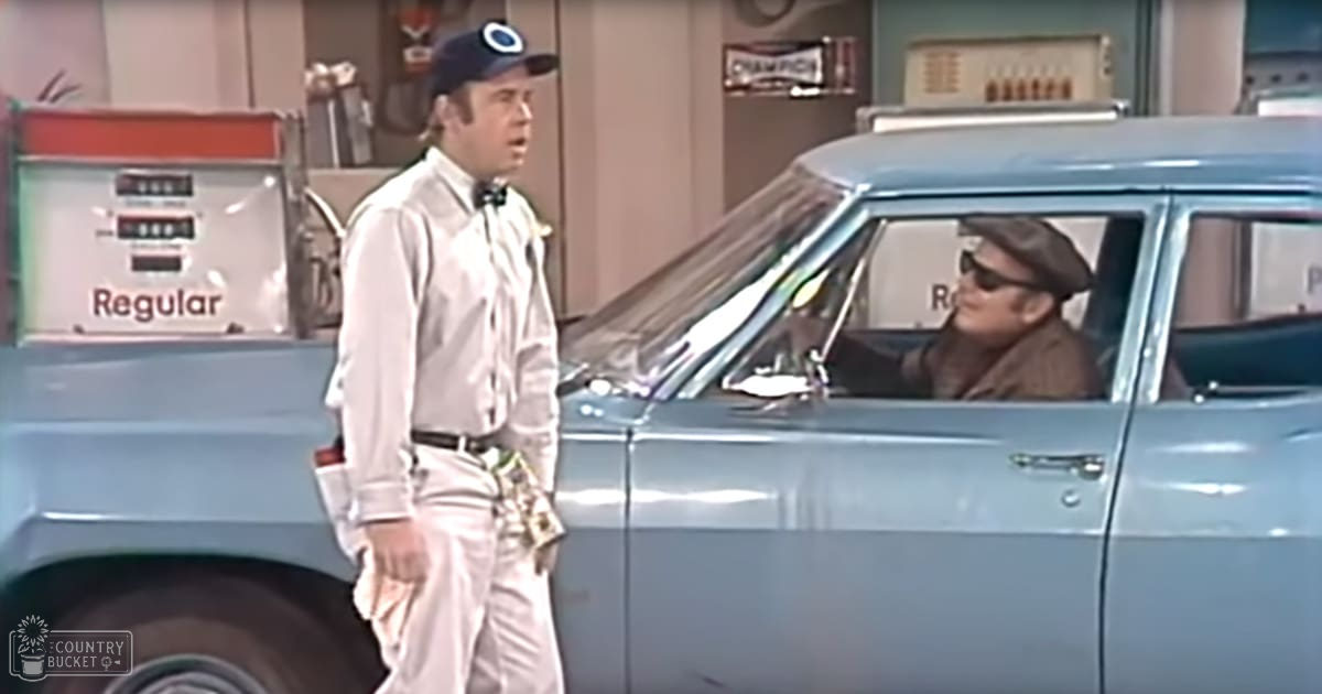 Adult male gas station attendant in a white shirt with rolled-up sleeves, bow tie, khaki pants, and cap holding keys; adult male driver inside an older model light blue sedan at a mid-20th century gas station.