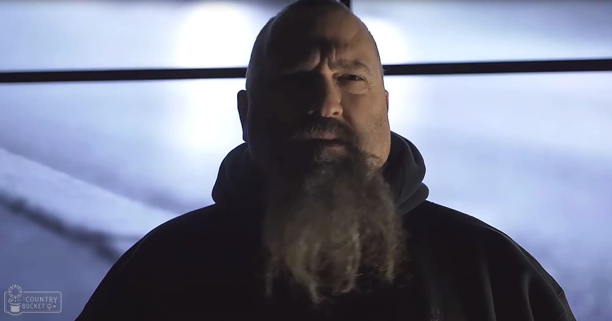 Older adult male wearing a dark-colored hoodie in a snowy outdoor setting with bright artificial lighting.