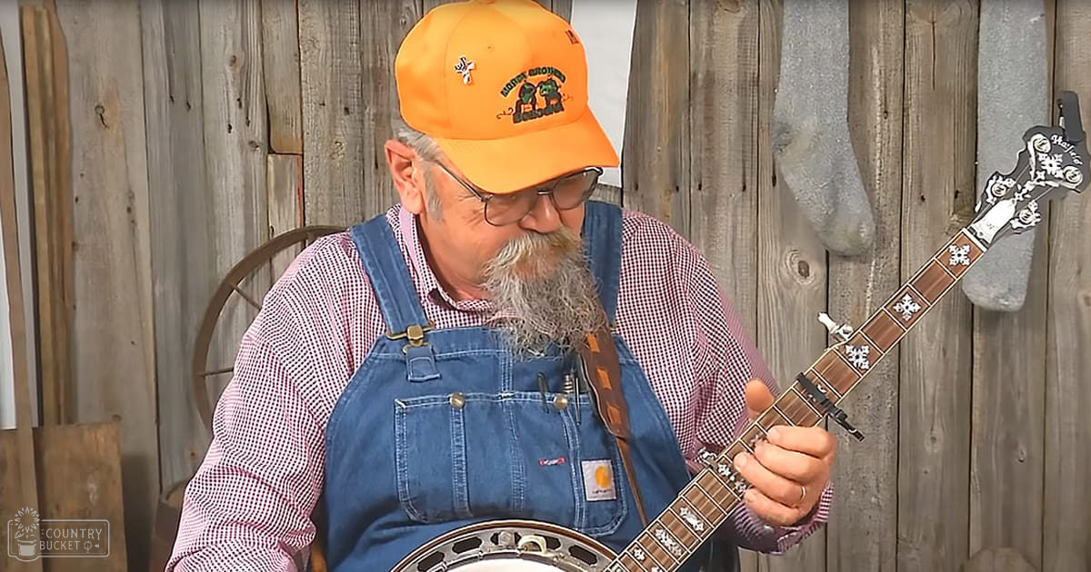 A middle-aged adult male playing a banjo indoors, wearing a red-and-white checkered shirt with blue denim overalls and an orange baseball cap.