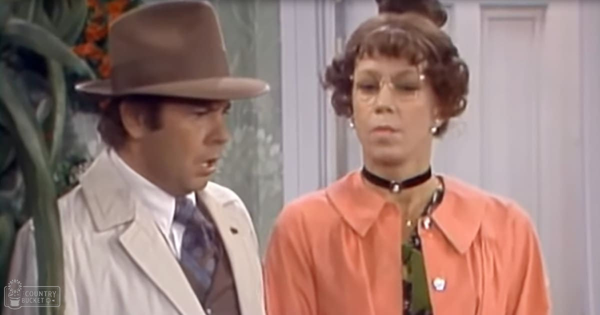 A woman in her middle years wearing an orange blouse with ruffled detail and a dark choker necklace stands indoors near decorative floral elements while a young adult male in a white trench coat, dark suit, patterned tie, and brown fedora hat is positioned nearby.