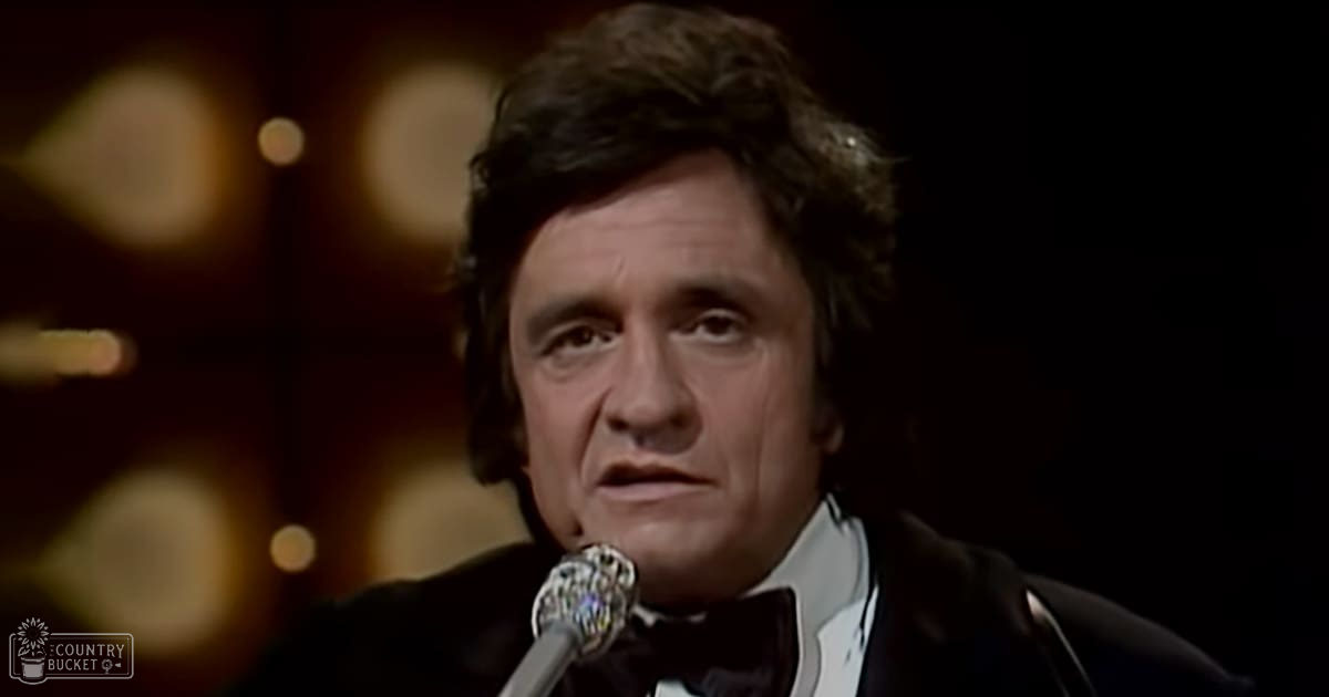 A middle-aged man wearing a black 1970s tuxedo with white shirt and bow tie, standing in an indoor venue with warm lighting possibly for a performance or event.