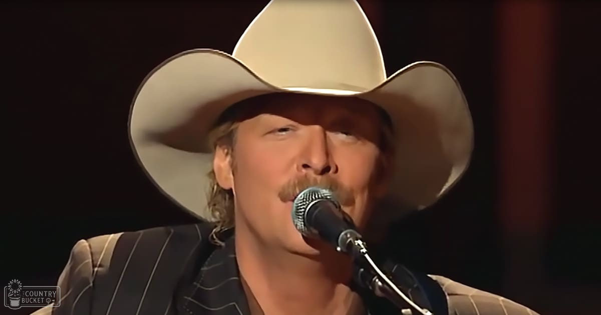 An adult male performer in a striped black and white pinstripe blazer with a collared shirt, holding a microphone while wearing a wide-brimmed cowboy hat, performing indoors at an evening concert or event.
