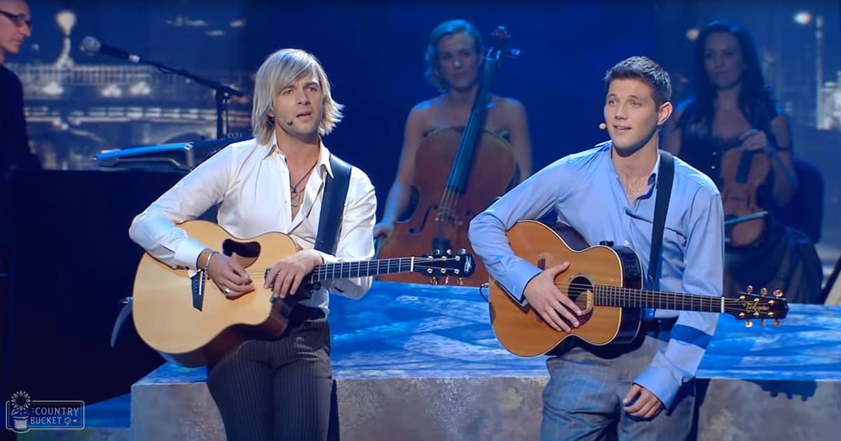 A musical performance scene with a young adult male in a white button-up shirt and dark pinstripe trousers on stage under evening lighting, with microphones and a cello in the background against an urban backdrop.