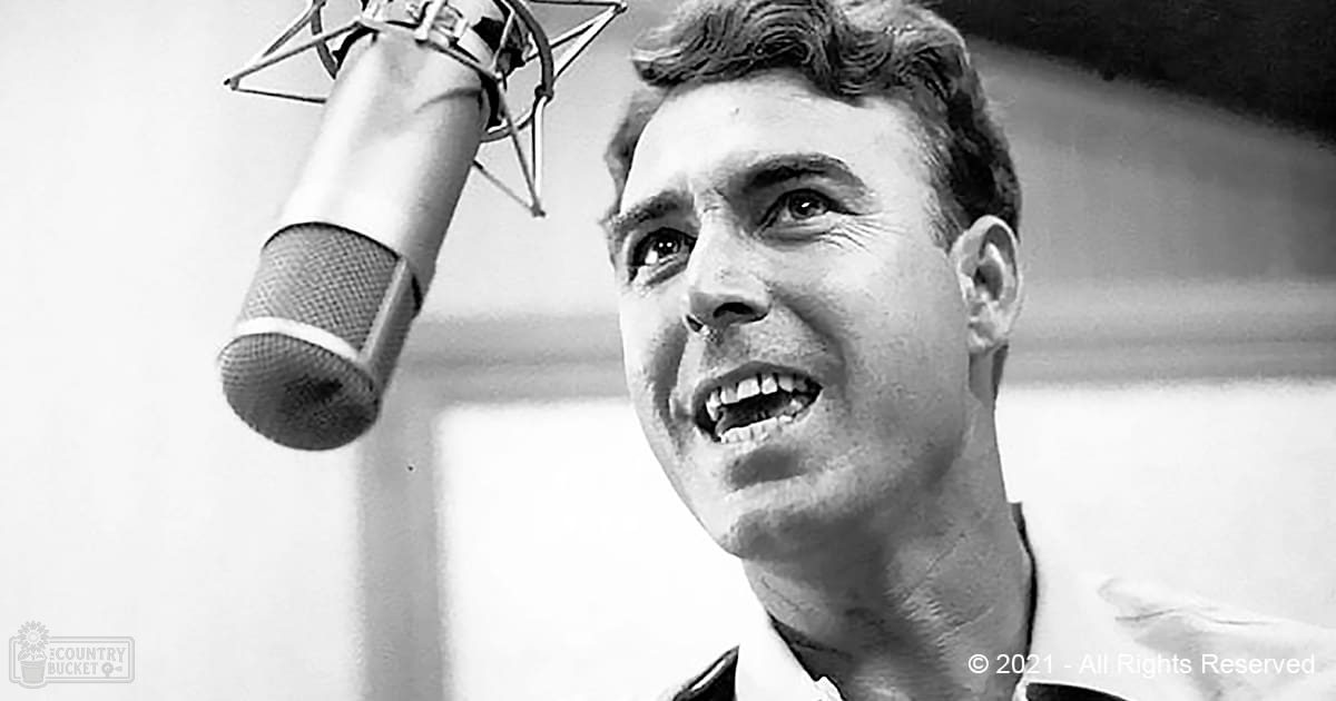 A young adult male in a recording studio wearing a formal-style shirt and jacket, with a professional microphone as the focal point against an indoor setup suggesting music production from the mid-20th century.