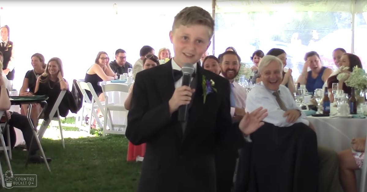Young boy in a tuxedo at an outdoor wedding reception under natural light with tables and chairs set up for guests.