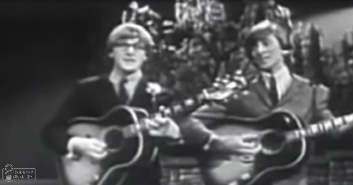A musical performance from the 1960s with two young adult male musicians in dark suits holding acoustic guitars on a stage, audience blurred in the background.