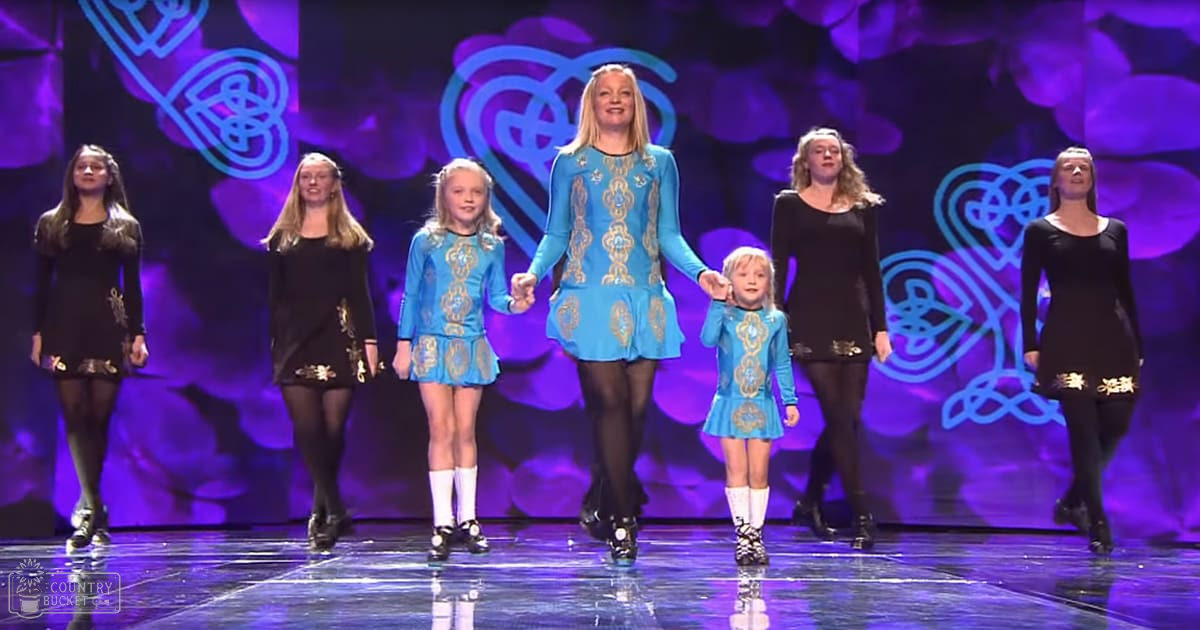 Group performing Irish dance with adults and children on stage in an indoor performance space during evening lighting.