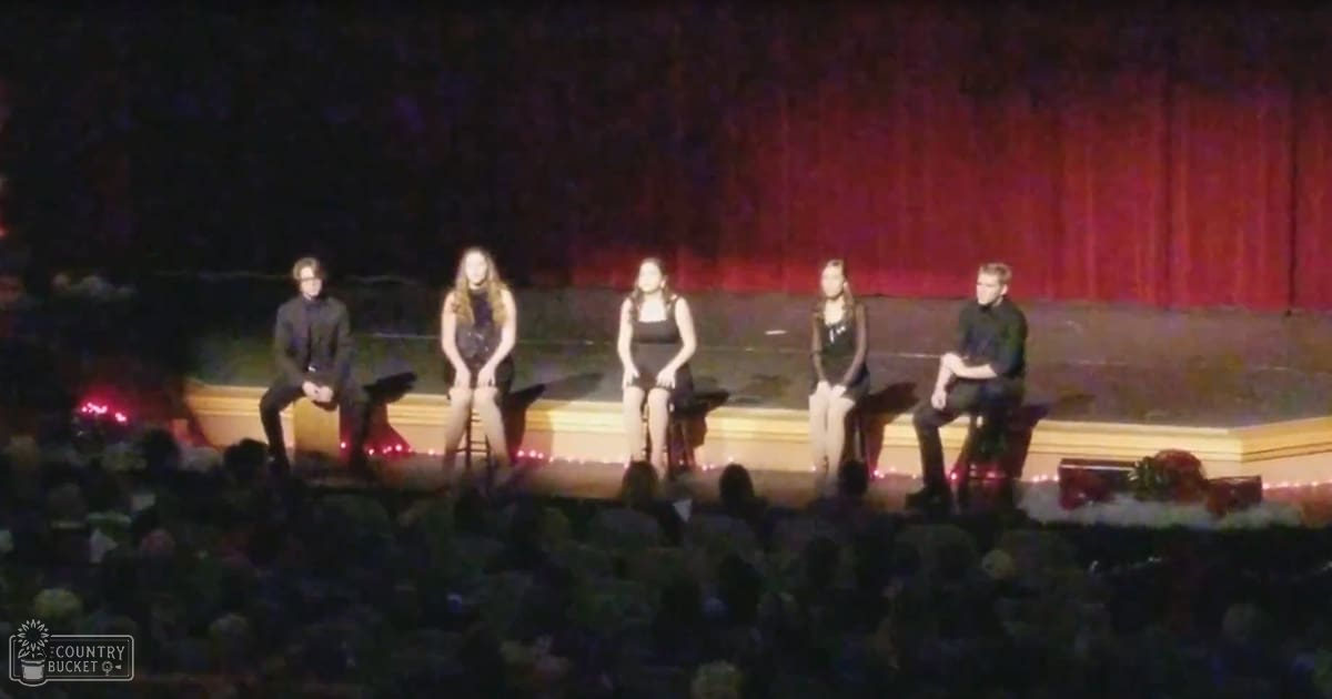 Panel discussion or Q&A session in an indoor auditorium with young adults wearing sleeveless black dresses and dark shirts; red curtain backdrop, dimly lit audience area.