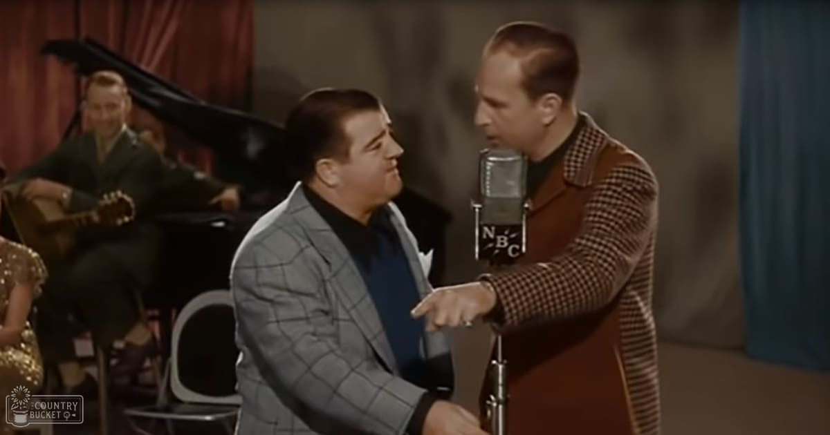 Musical performance in a studio setting with an adult male singer wearing a gray plaid suit, seated on stage, and another middle-aged to older adult male standing nearby in a brown checkered jacket over a black turtleneck sweater. Microphone labeled 'NBC' is prominently featured.