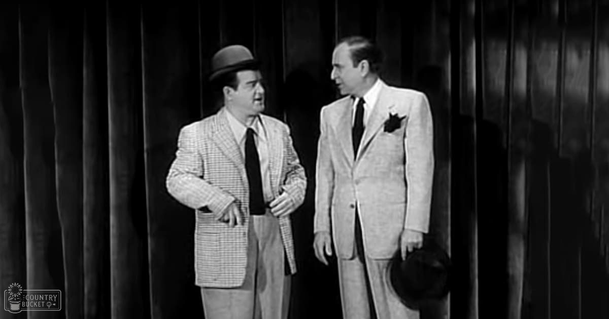 Two middle-aged men in checkered suits stand against a dark curtain backdrop, suggesting an indoor performance or meeting setting with dramatic lighting.