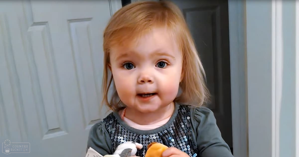A toddler wearing a dark green long-sleeved shirt with a shiny black and silver sequined bib, standing indoors near a white door. The lighting suggests daytime.