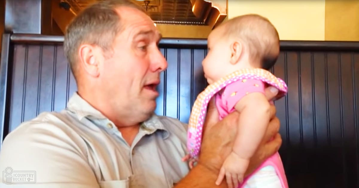 A middle-aged male holds an infant wearing a pink and white polka-dotted outfit in what seems to be a restaurant with dark wooden paneling and artificial lighting.