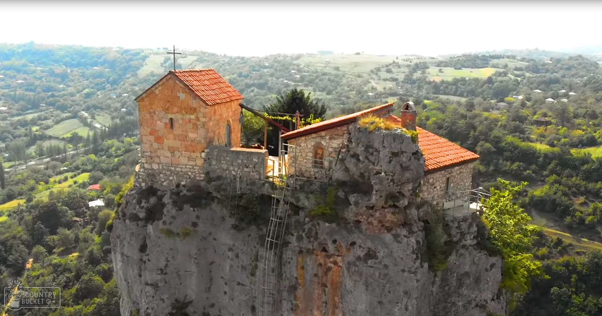 A small stone church with red-tiled roofs and crosses on a rocky outcrop amidst lush green rolling hills under natural daylight.