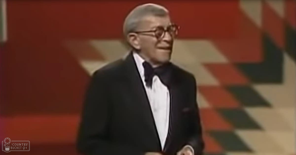 An older adult male in a tuxedo, wearing a bow tie and rectangular-framed eyeglasses, set against a background with geometric patterns and warm tones.