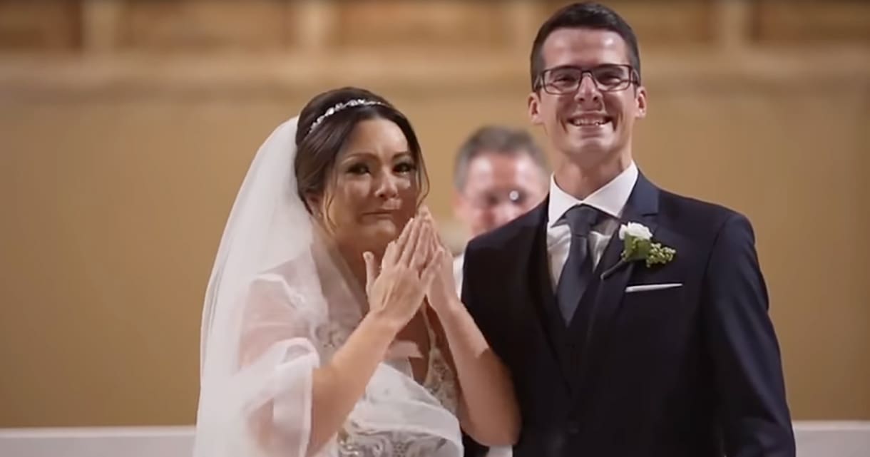 A bride and groom at their wedding ceremony indoors with a warm-toned wall, possibly a wooden ceiling or paneling, and natural daylight or indoor lighting.
