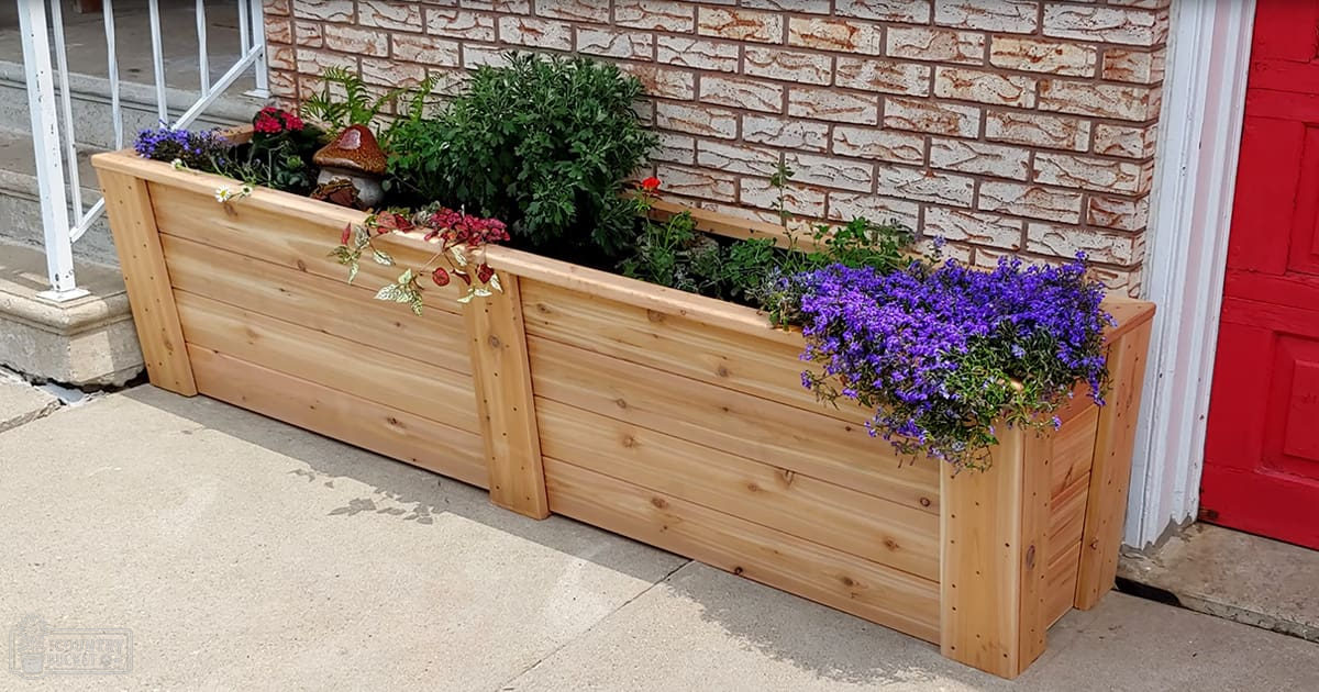 Two wooden planter boxes filled with various plants and flowers in front of a house with a brick facade, red door, and white railing.