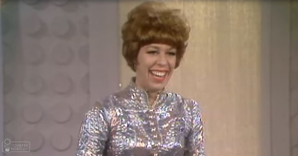 A woman wearing a silver sequined high-necked, long-sleeved form-fitting dress indoors with patterned wallpaper in the background.