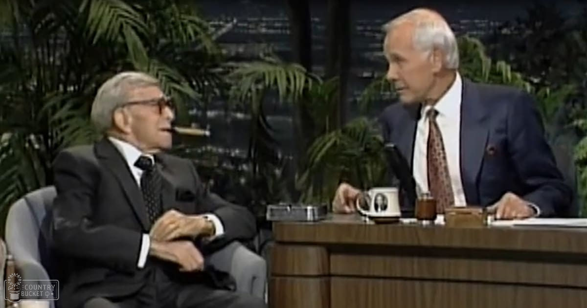 Two older adult males in a talk show setting, discussing, surrounded by greenery and cityscape views.