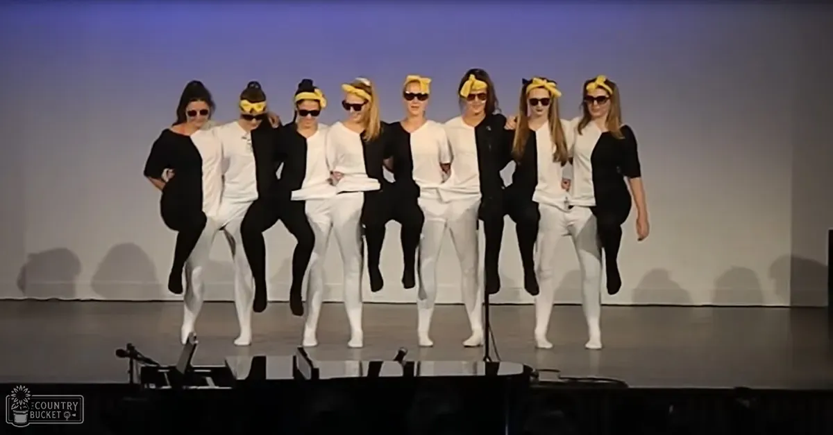 A group of young adult females in loose-fitting white and black outfits perform on stage under artificial lighting.