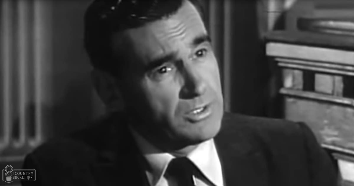 A middle-aged adult male wearing a dark suit jacket, white shirt, and tie in an indoor setting with neutral lighting.