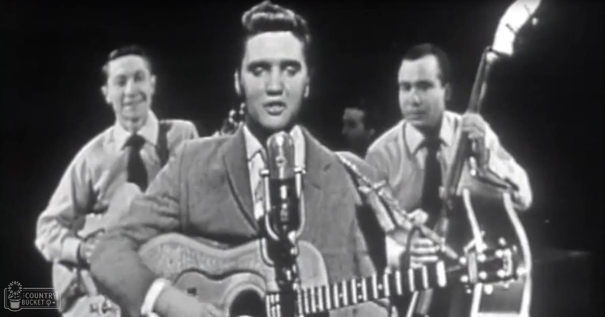A young adult male performer wearing a suit jacket and tie stands on stage with two musicians behind him, all dressed in light-colored shirts and dark ties, under artificial indoor lighting.