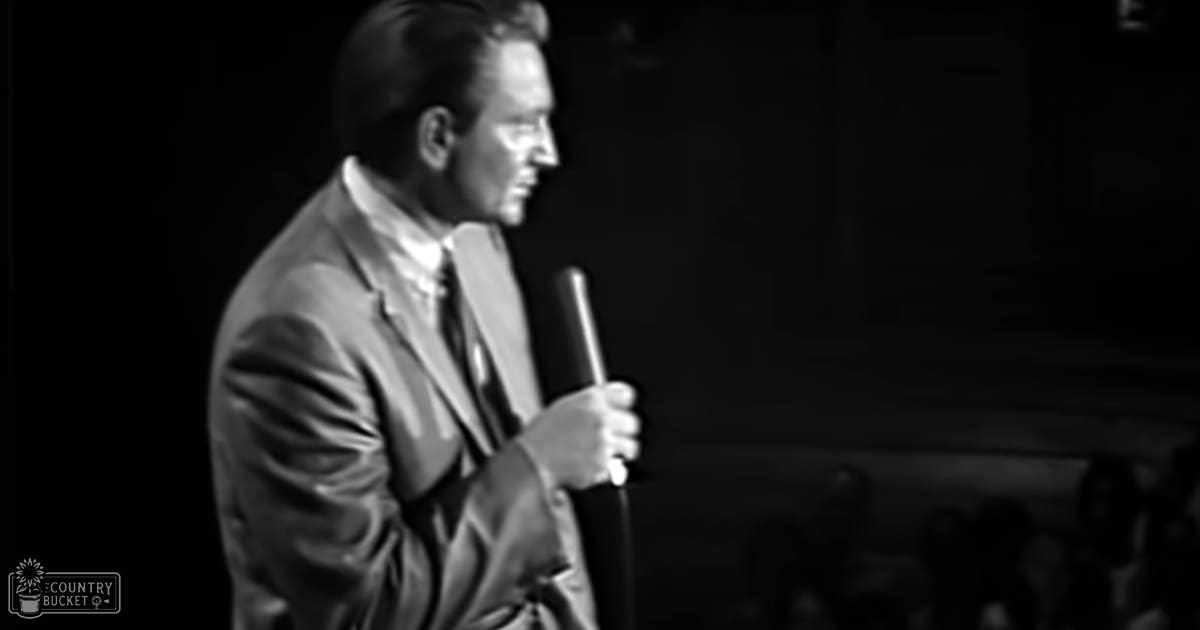 A middle-aged man performing stand-up comedy indoors with a dark, stage-like background.
