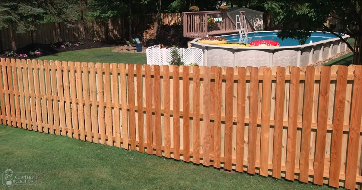 A residential backyard with a wooden picket fence, an above-ground swimming pool, inflatable rings, white lattice privacy screen, green grass, and trees in the background.