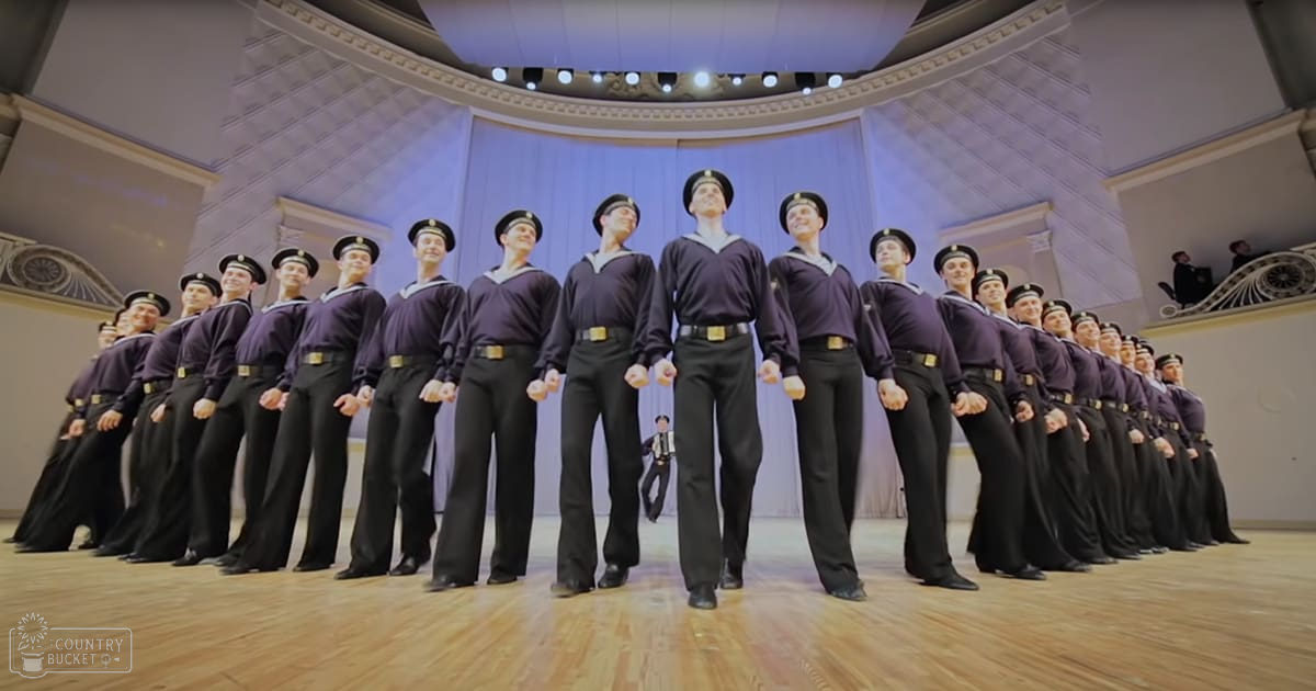 Young adult to adult men in dark navy blue sailor-style uniforms with white trim, black belts, and black hats, performing indoors within an ornate auditorium or theater at night.