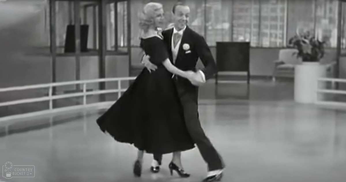 A couple dancing in a dance studio or ballroom, dressed formally with black attire and a white shirt, surrounded by indoor furniture and greenery.