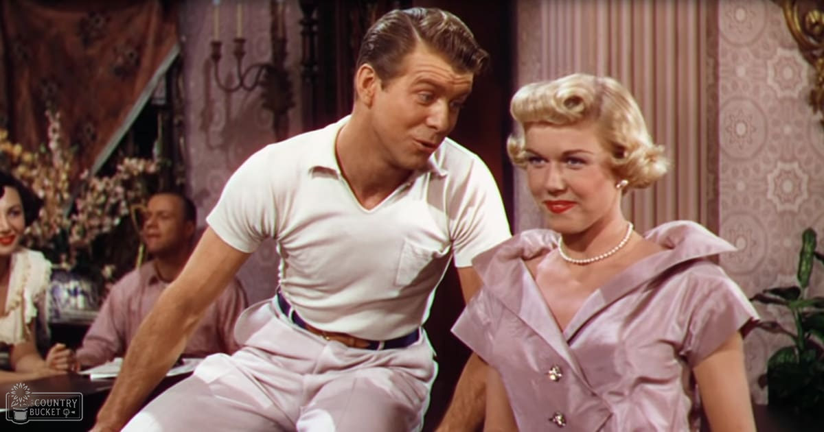 Two adults, a man wearing a white short-sleeved shirt with pocket details and light-colored trousers, standing indoors with ornate mid-20th-century decor; the woman wears a light purple dress with pearl jewelry in an indoor social setting.