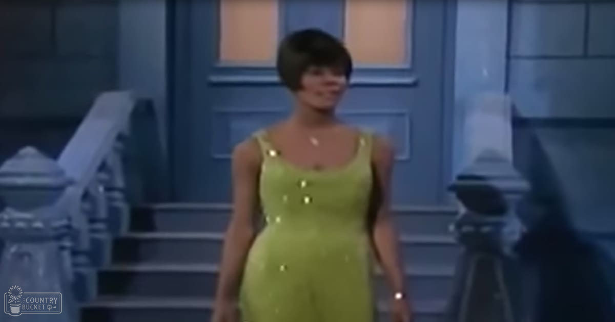A young adult female wearing a bright green sleeveless dress with a fitted bodice and scoop neckline stands indoors near a staircase with ornate railings and blue paneling, suggesting a home environment.