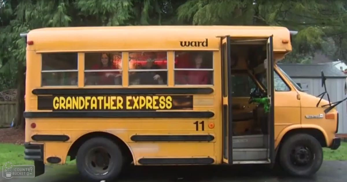 A yellow school bus labeled 'Grandfather Express' is parked outdoors in a residential setting, surrounded by trees and a small shed-like structure under natural daylight.