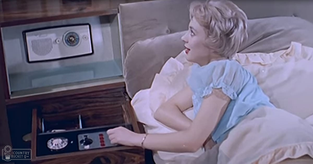 A young adult female wearing a light blue nightgown with ruffled edges and short sleeves in a bedroom setting with vintage furniture.