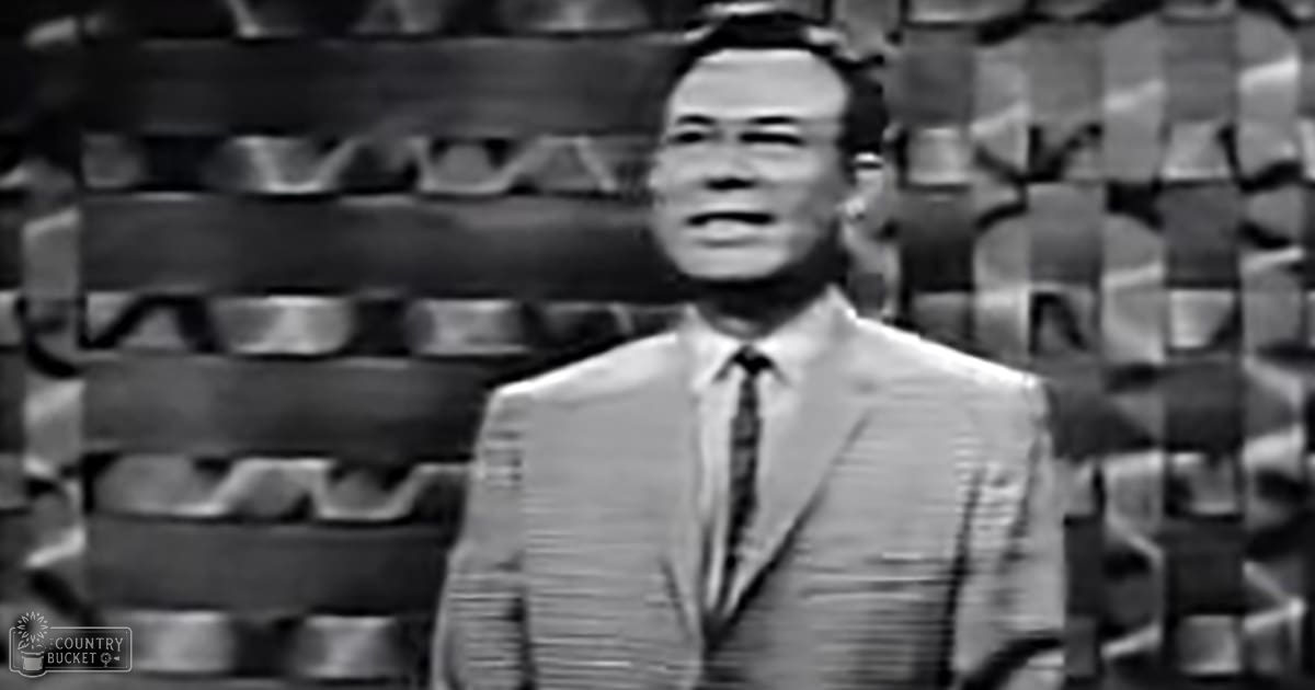 Middle-aged male in a tailored suit jacket with a collared shirt, light-colored fabric with subtle stripes or patterns, likely from an earlier era; indoors, possibly a television studio set with geometric patterned backdrop.
