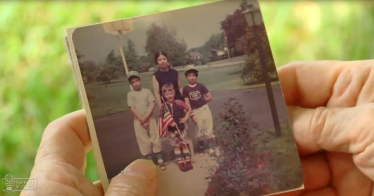 Group photo taken in an outdoor residential area with children and adults, basketball hoop visible in the background, American flag held by one of the children.