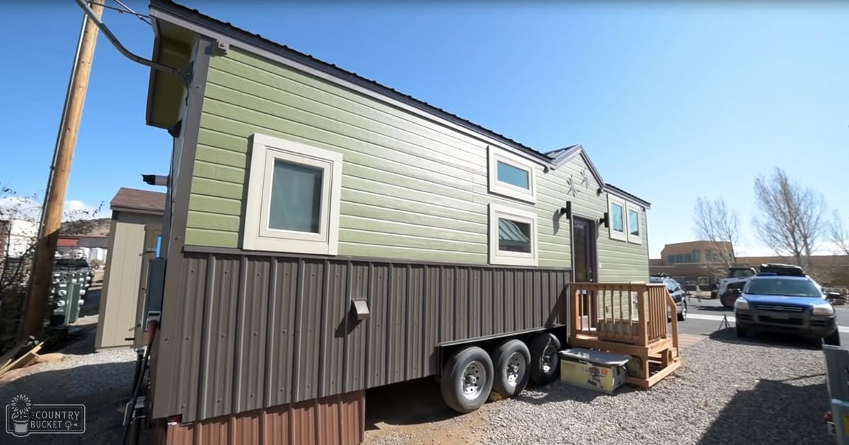 A small trailer with wheels supports a tiny house on an outdoor wooden deck, surrounded by bare trees and a parked car under clear blue skies during late fall or winter.