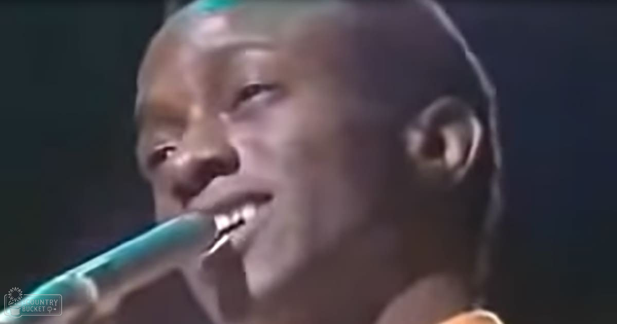 A young adult male singing into a microphone at an indoor venue under artificial stage lights, wearing an orange tank top with teal accents.