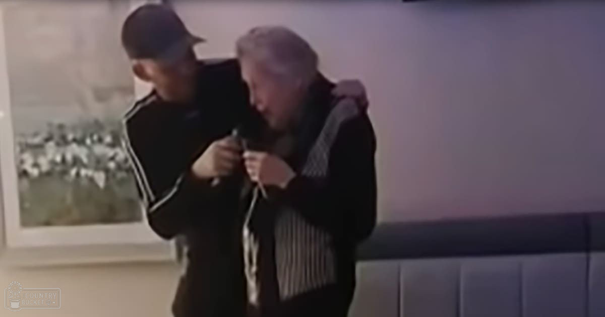 An older adult male wearing a patterned scarf and modern jacket indoors alongside a younger adult in a dark cap and striped shirt.