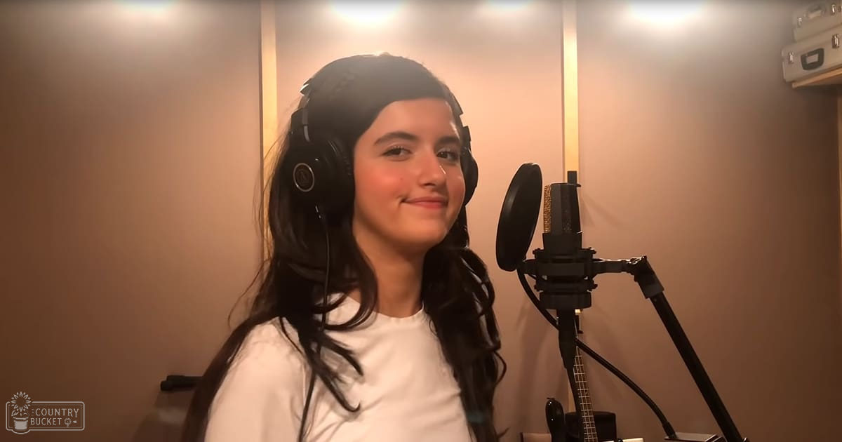 A young adult female in a recording studio, wearing a white t-shirt with headphones, surrounded by acoustic foam panels and a microphone setup.