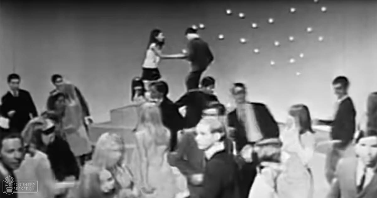 An indoor dance performance with young adults wearing mid-20th century formal attire on a stage with circular light patterns, likely evening or night time, muted black and white tones.