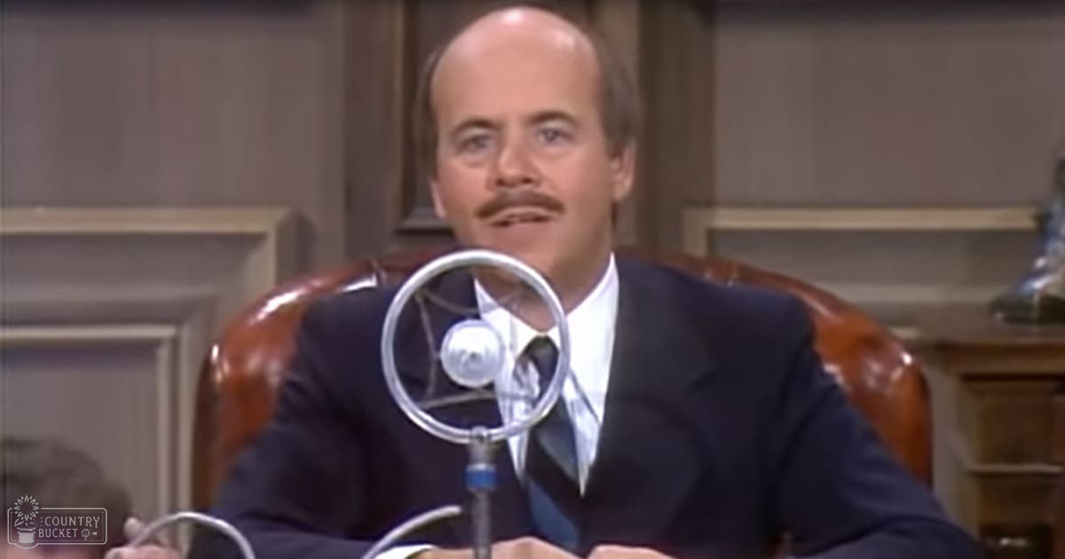 A middle-aged to older adult male in a formal setting, wearing a dark suit jacket and white dress shirt with a dark tie, seated at a table with a microphone, surrounded by wood paneling and formal decor.
