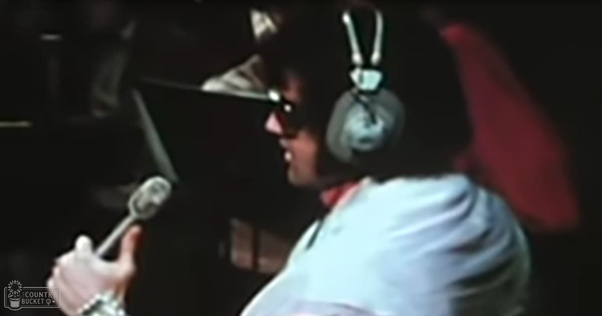 Adult male singer wearing headphones, white t-shirt, and sunglasses, passionately singing into a microphone in a recording studio with red and black elements.