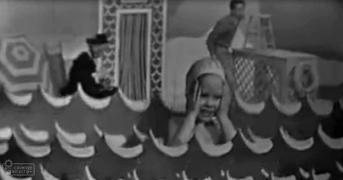 A toddler wearing a light-colored dress indoors alongside adults dressed formally from the early-to-mid 20th century in what appears to be a theatrical or entertainment venue.