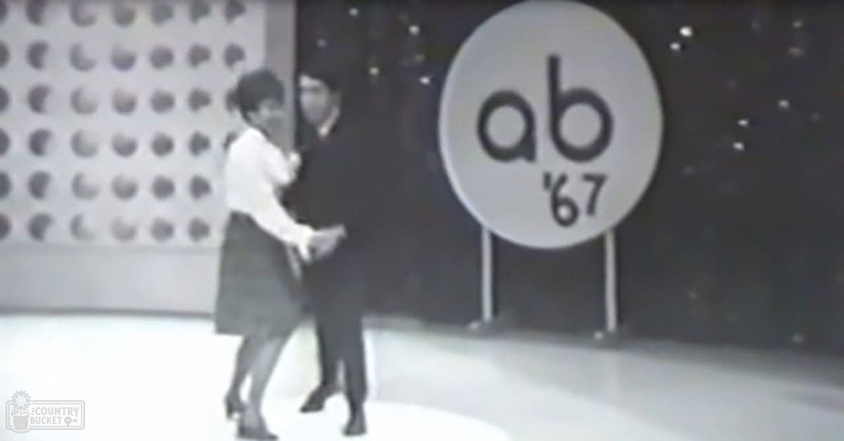 A young adult female in a white blouse with a collar dances alongside an adult male in a dark suit at AB '67, surrounded by 1960s decor and patterned walls.