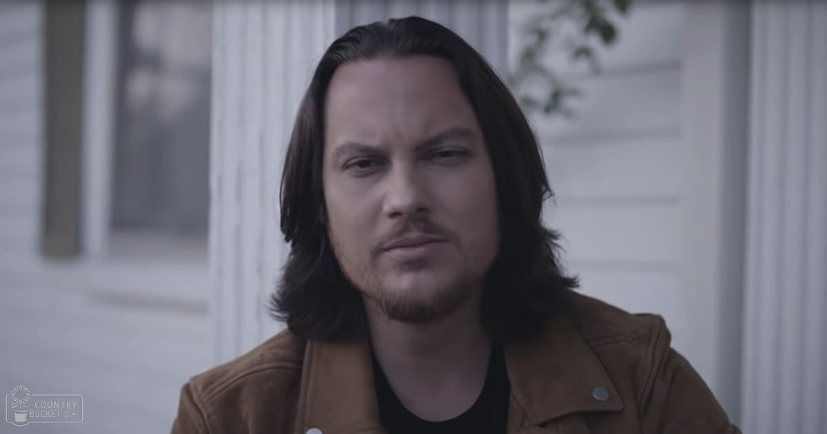 Young adult male in brown leather jacket outdoors with white-sided building and spring-greenery backdrop, late afternoon light.